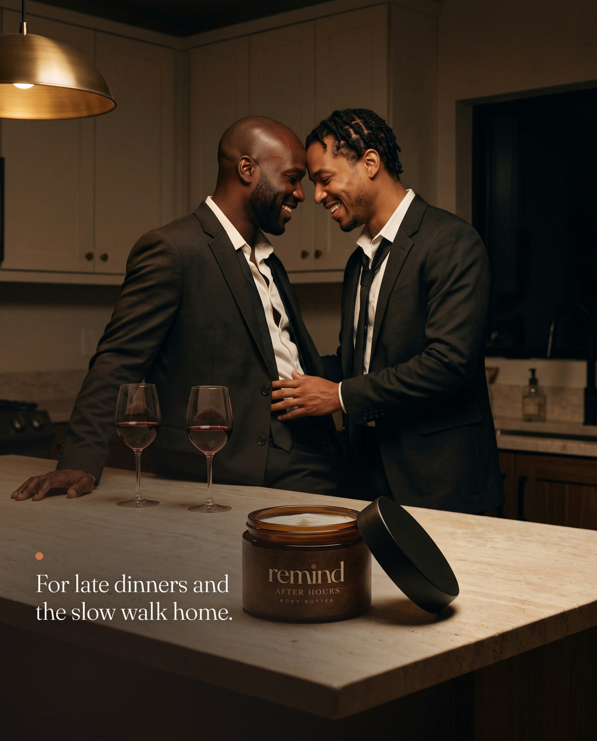 Two men in loosened suits sharing a close moment in the kitchen after dinner; an open jar of REMIND AFTER HOURS body butter on the island. Caption: For late dinners and the slow walk home.