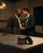 Two men in loosened suits sharing a close moment in the kitchen after dinner; an open jar of REMIND AFTER HOURS body butter on the island. Caption: For late dinners and the slow walk home.