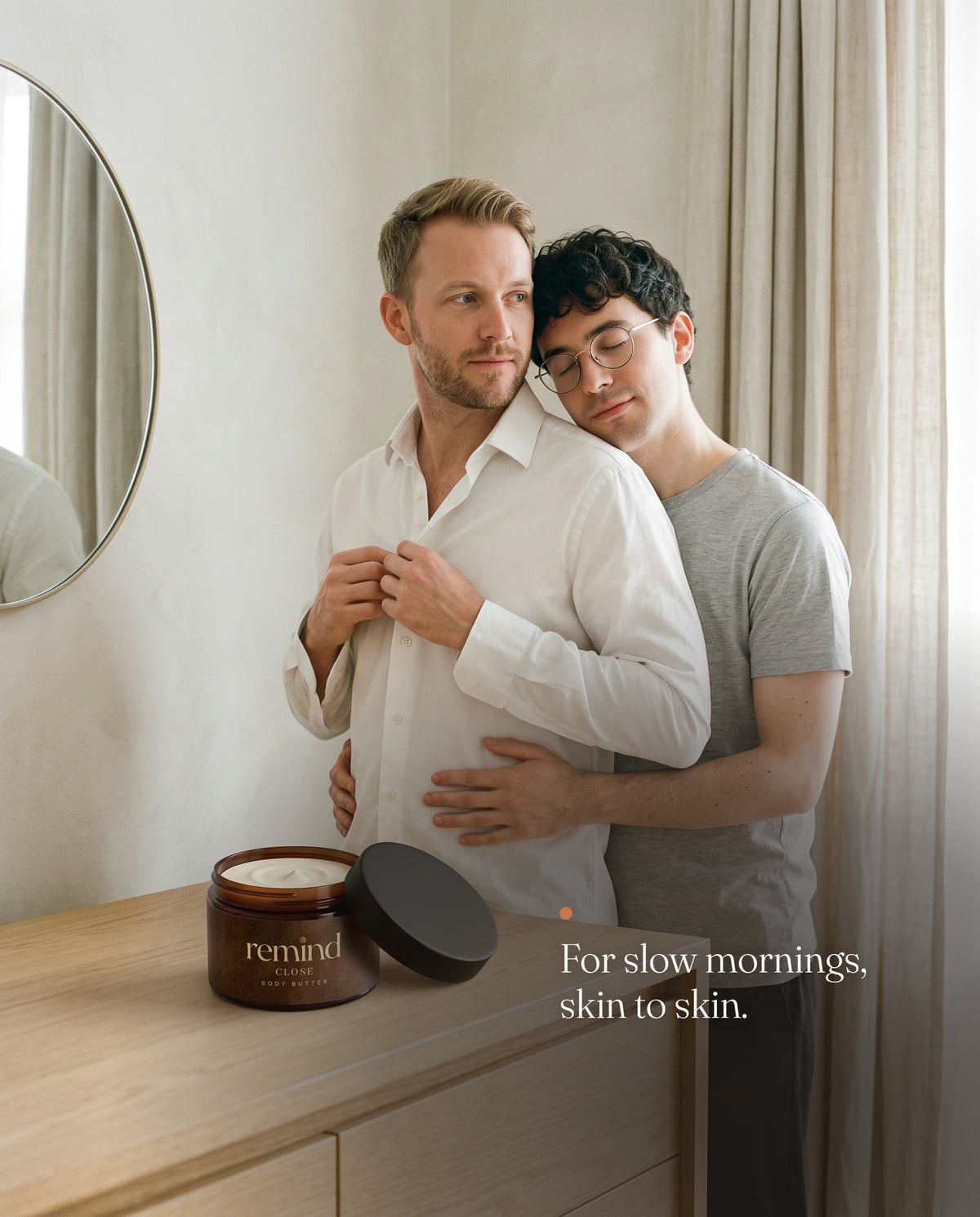 A man buttons his white shirt while his partner rests his cheek on his shoulder; an open jar of REMIND CLOSE body butter on the dresser. Caption: For slow mornings, skin to skin.