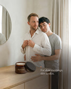 A man buttons his white shirt while his partner rests his cheek on his shoulder; an open jar of REMIND CLOSE body butter on the dresser. Caption: For slow mornings, skin to skin.