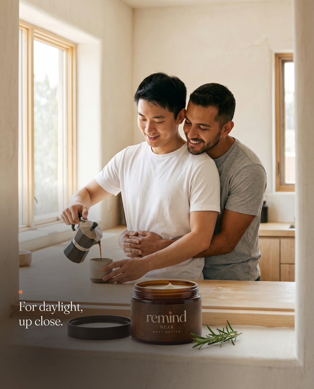 One man hugs another from behind as he pours coffee on a sunny morning; an open jar of REMIND NEAR body butter on the windowsill. Caption: For daylight, up close.
