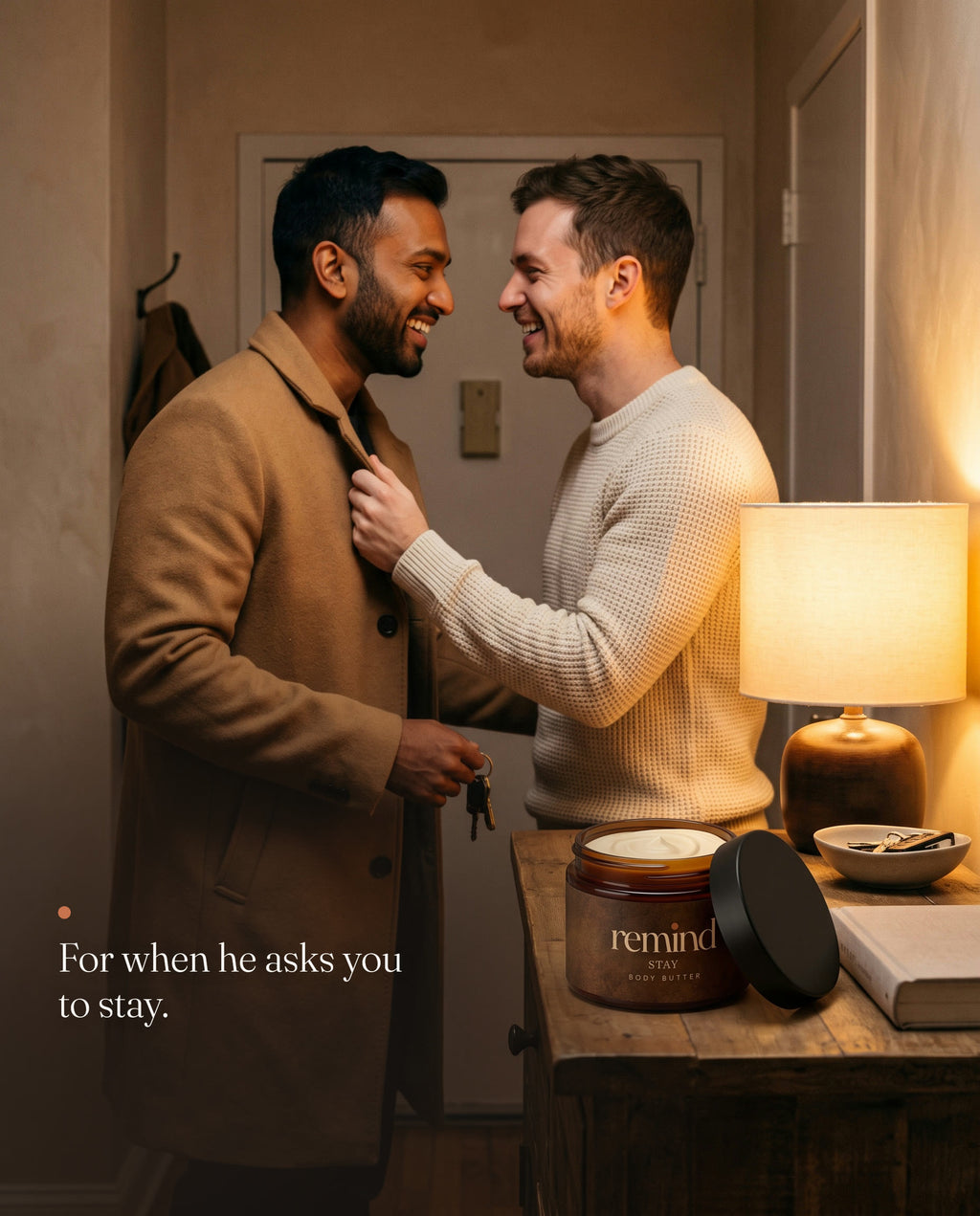 Two men laughing in an apartment entryway, one holding the other back by his coat lapel; an open jar of REMIND STAY body butter on the console. Caption: For when he asks you to stay.