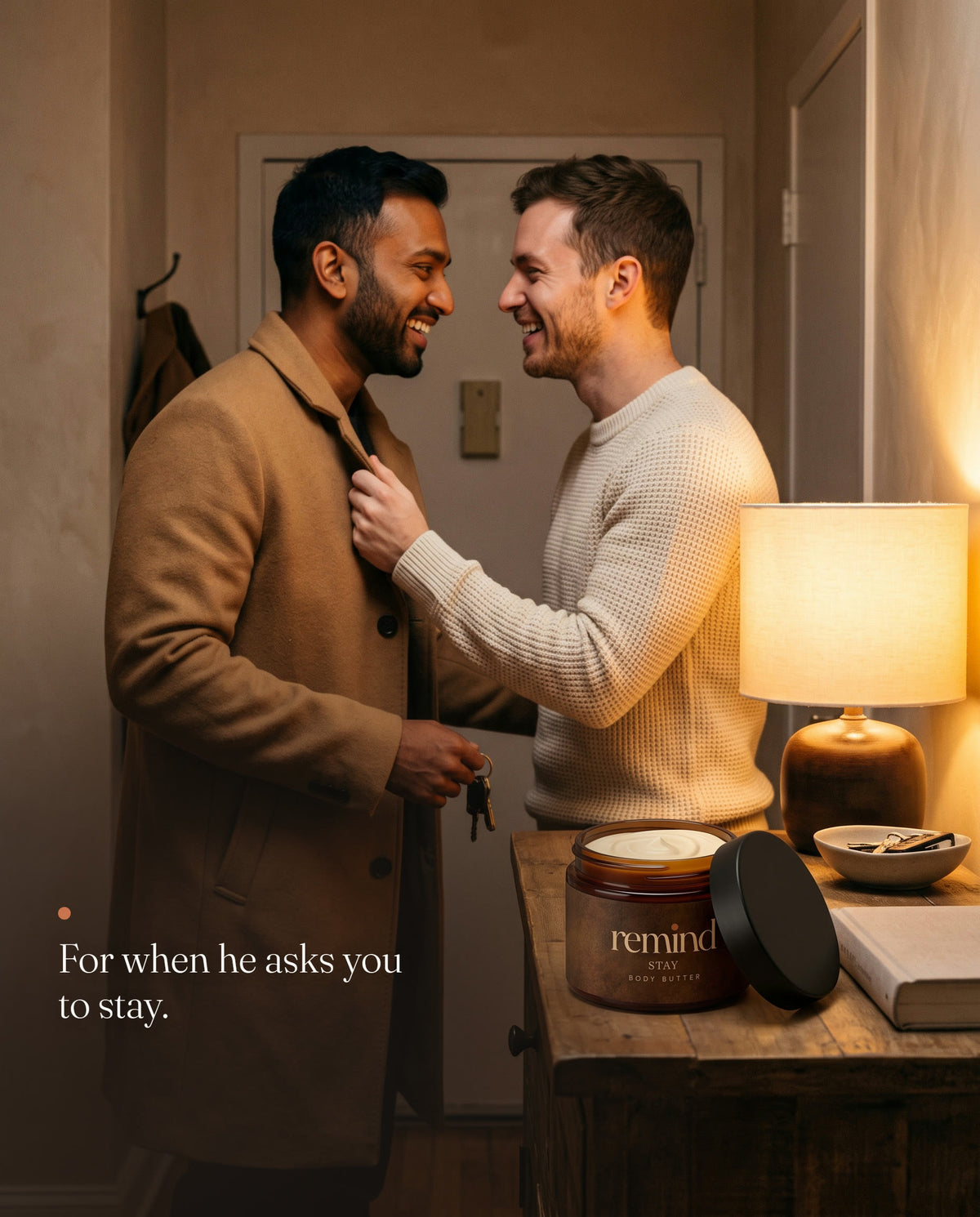 Two men laughing in an apartment entryway, one holding the other back by his coat lapel; an open jar of REMIND STAY body butter on the console. Caption: For when he asks you to stay.